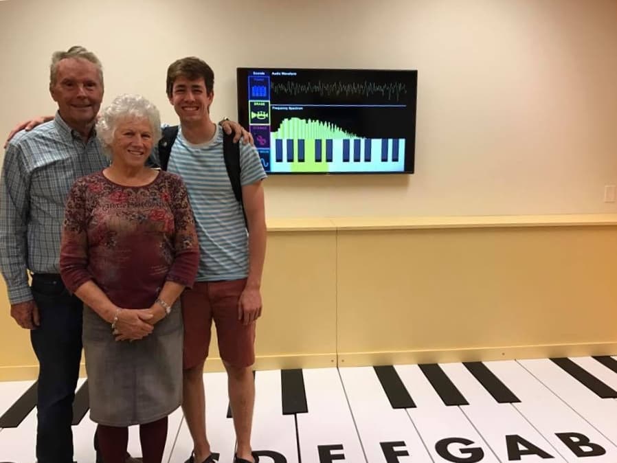 My grandparents and I standing in front of a large piano keyboard on the ground with my software running on a screen attached to the wall.