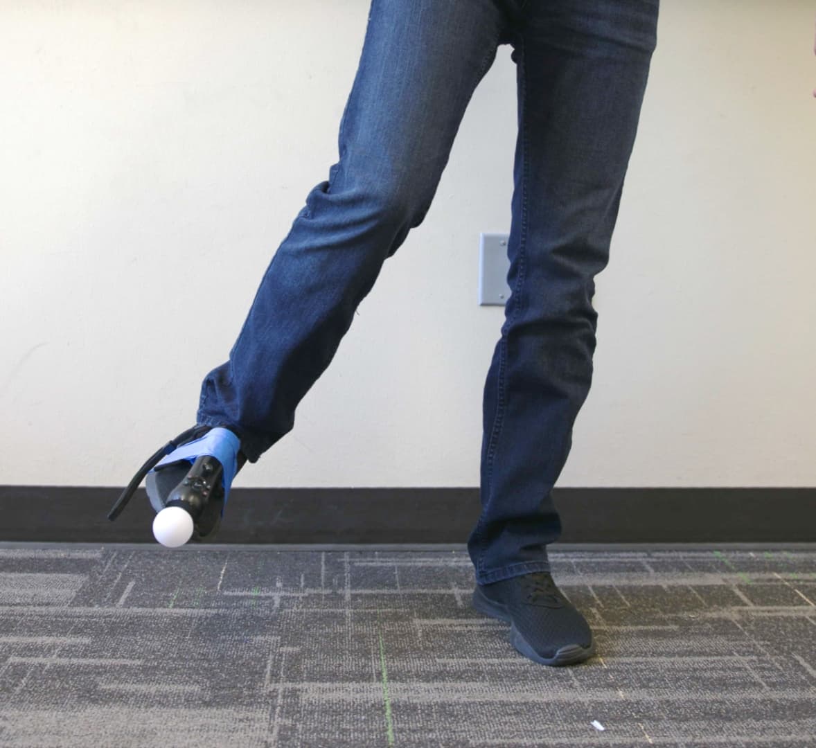 A foot wearing a shoe with a PlayStation Move motion controller taped to it. The leg is being swung to the user's right.