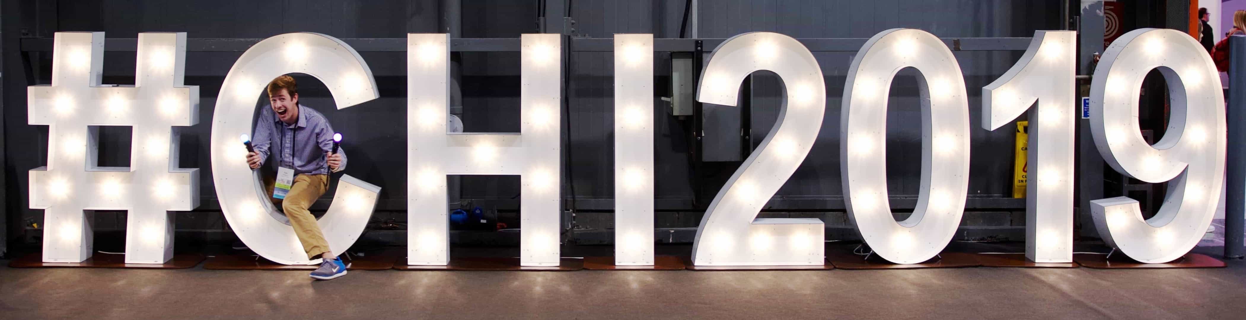 Photo of Tim standing in front of a sign that says #CHI2019 while holding the PlayStation Move controllers.